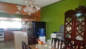 A dining area with a wooden table, chairs, and a chandelier. There is a kitchen in the background with a water dispenser and cabinets. - Victoria's Place (Portmore)