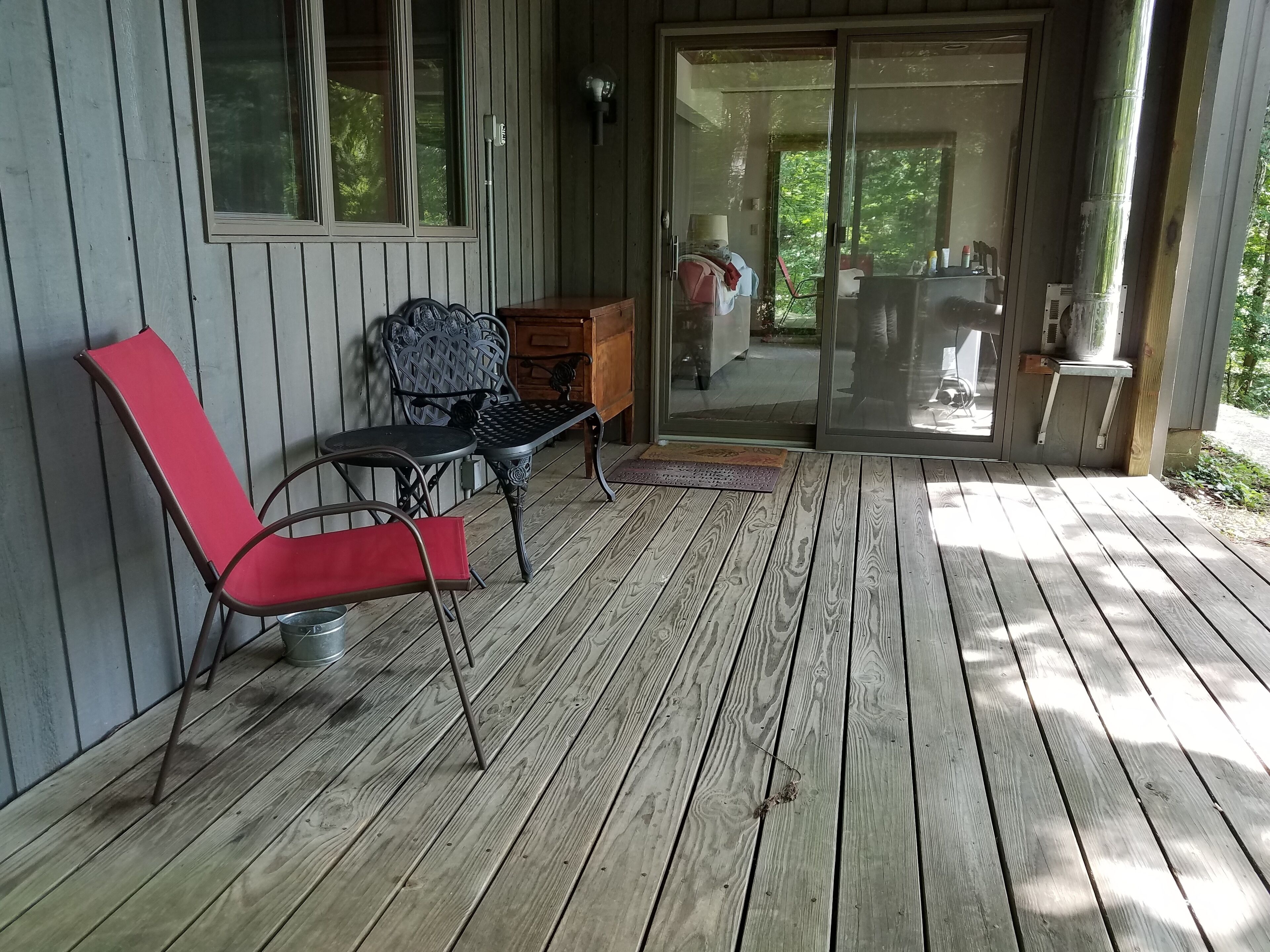Une terrasse en bois avec deux chaises et une table, une porte-fenêtre coulissante donnant sur un patio, et un petit meuble en bois.