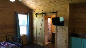 Una habitación de cabaña de madera con una cama, un televisor, una ventana y una puerta que da a otra habitación.