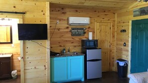 Interior de una cabaña de madera con cocina, televisor montado, microondas, refrigerador y un cesto de basura.