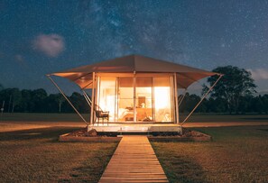Escena nocturna iluminada por estrellas, con una pequeña estructura al aire libre que tiene una terraza de madera que conduce a una entrada con puerta de vidrio.