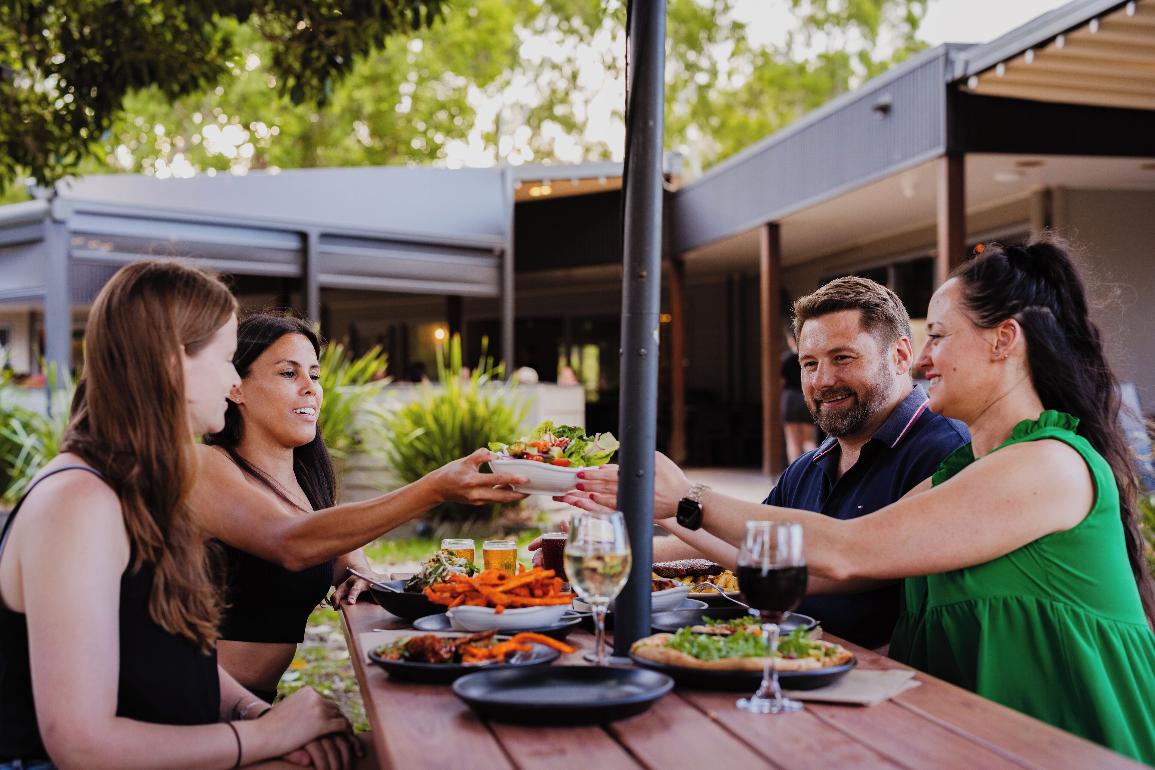 Un grupo de personas cenando al aire libre en una mesa de madera con diversos platos y bebidas.