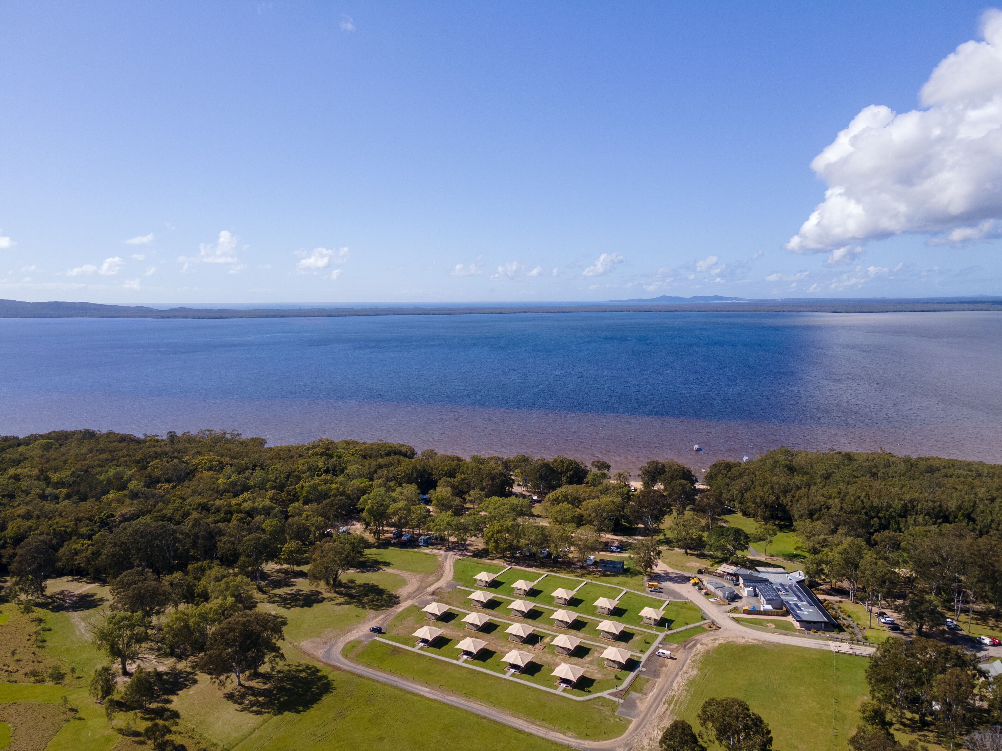 Vista aérea de un camping con tiendas de campaña dispuestas en filas, un camino y una masa de agua al fondo.