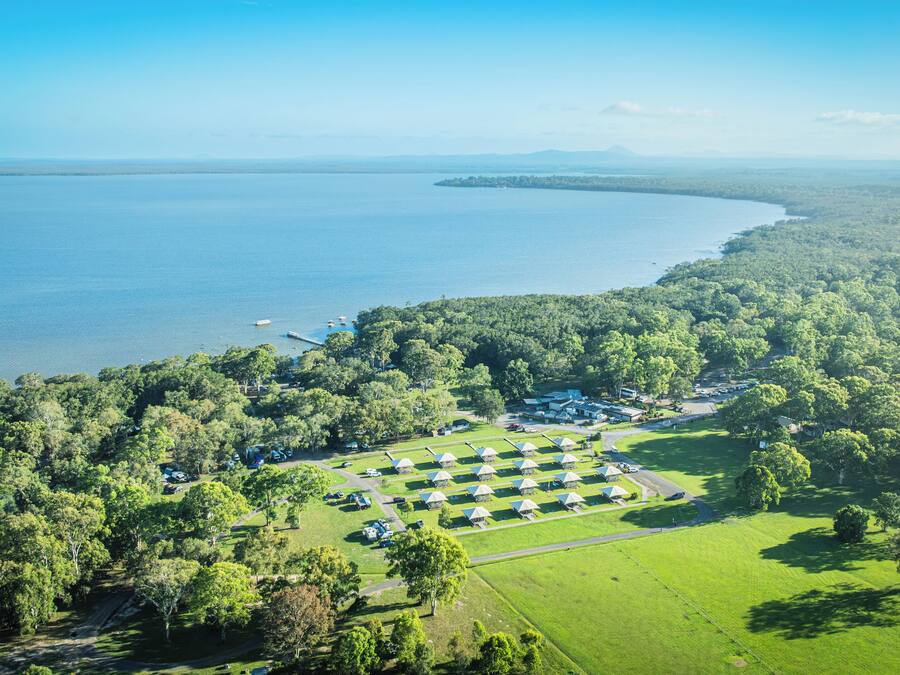 An aerial view of a campground with yurts arranged in rows, surrounded by dense forest, with a large body of water and distant mountains.