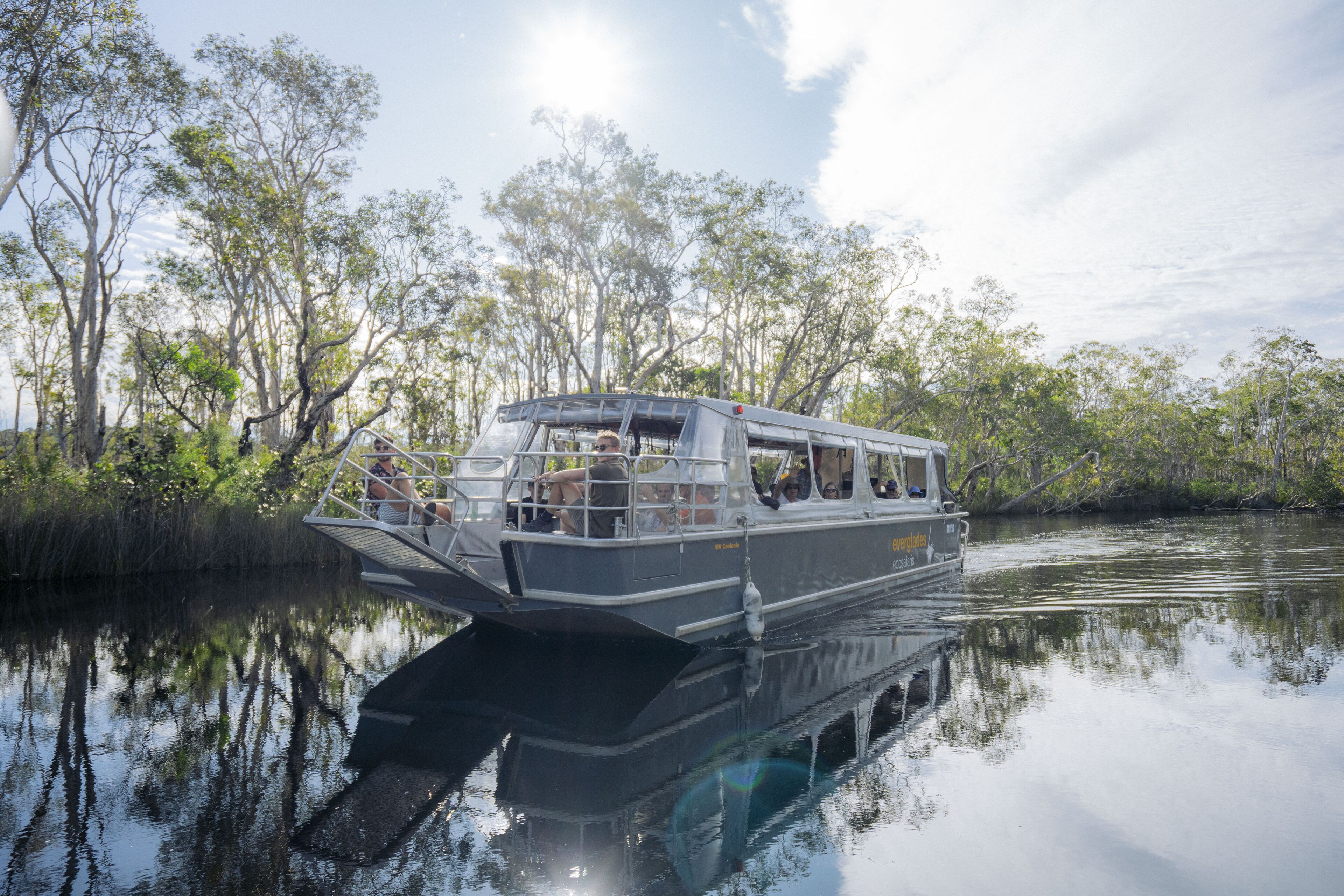 A riverboat with a covered deck, passengers, and a calm water body surrounded by trees.