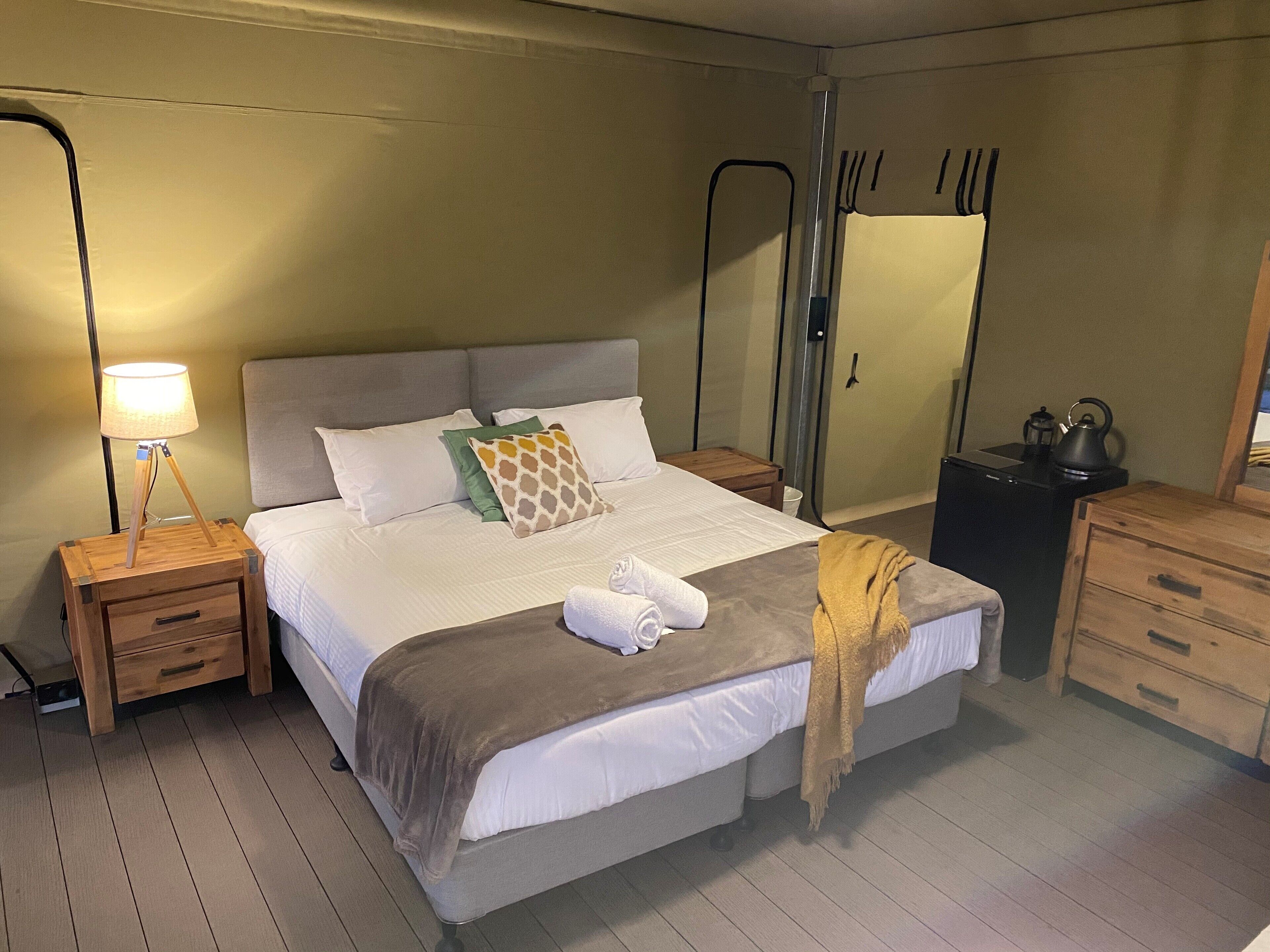 A neatly made bed with pillows and a blanket, a bedside lamp, a wooden nightstand with a drawer, a wooden dresser, and a mirror on the wall.