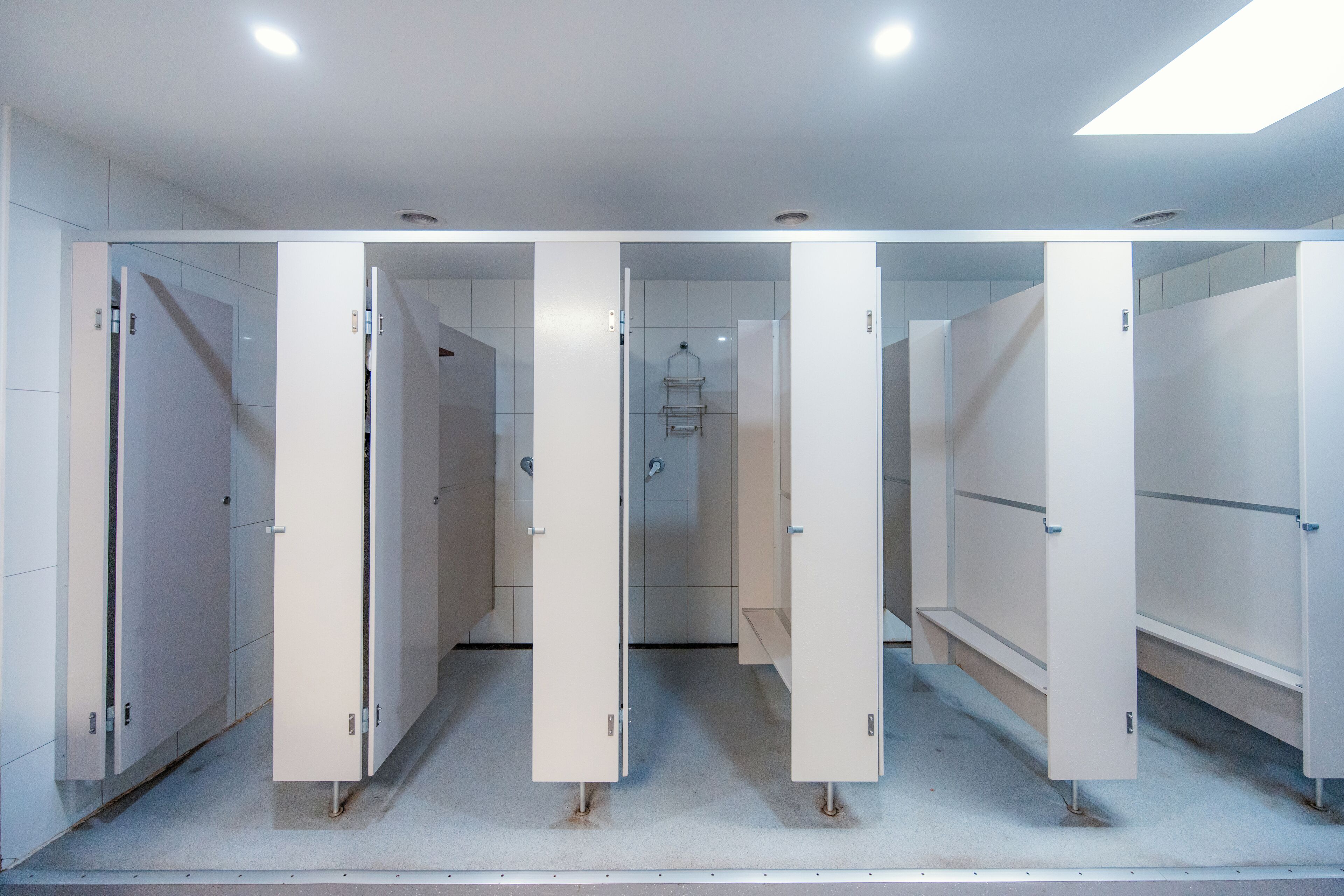 A row of white restroom stalls with a tiled floor and a skylight above.
