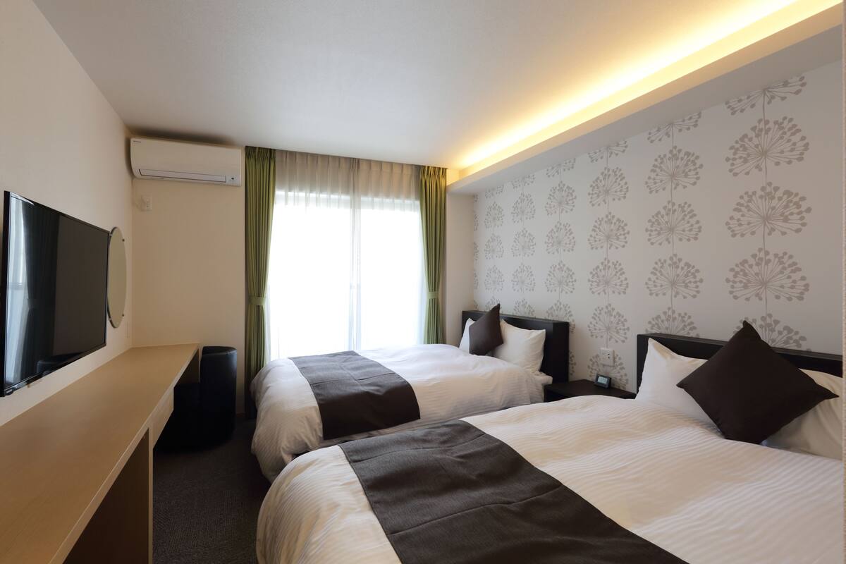 A hotel room with two beds, a TV, an air conditioner, and a window with curtains.
