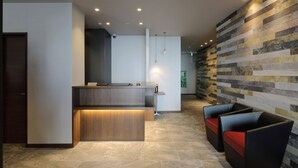 A modern reception area with a wooden reception desk, a seating area with cushioned chairs, and a wall with a mosaic tile design. - Hotel Promote Hiroshima (Hiroshima)