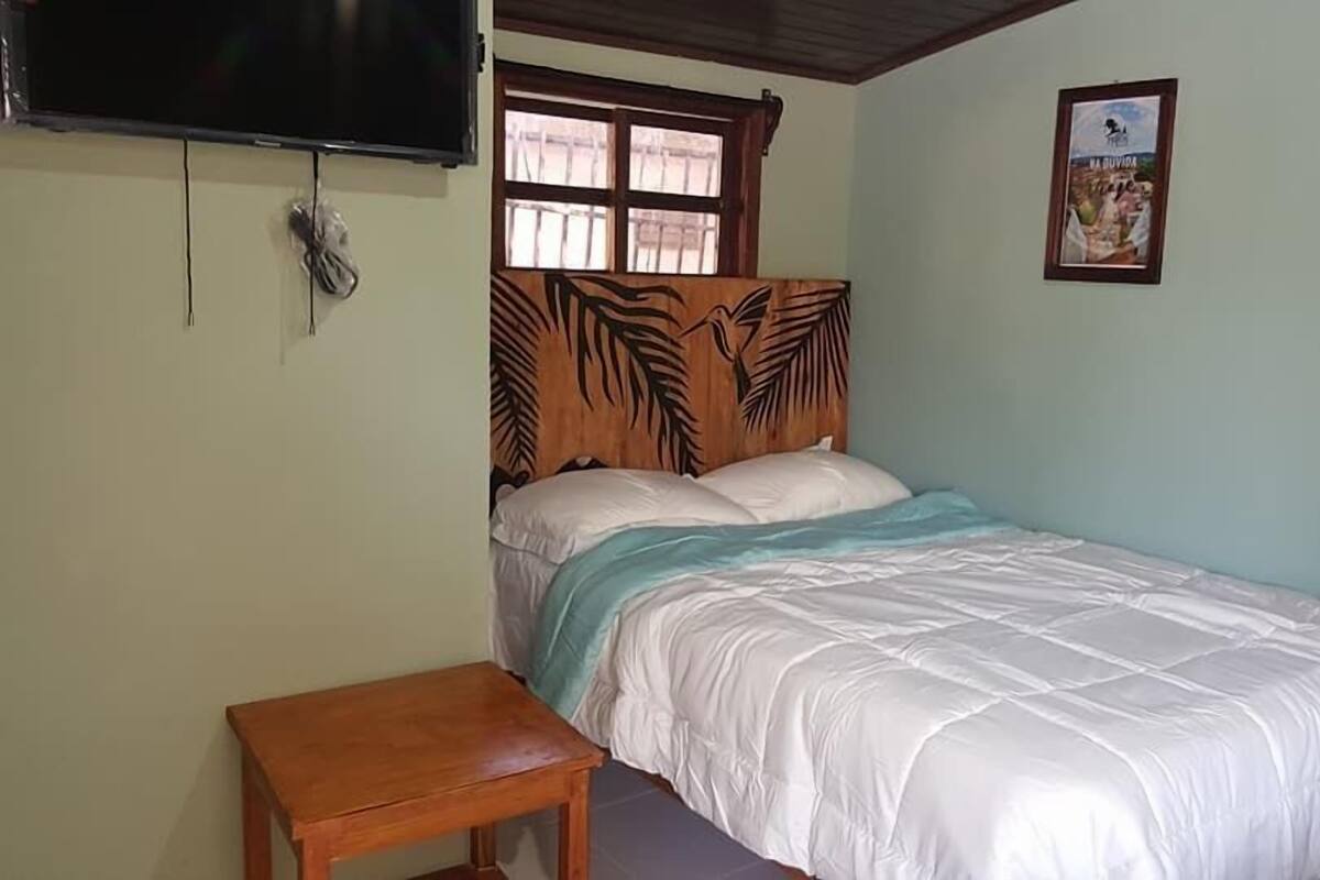 A bedroom with a bed, a wooden headboard, a small wooden table, a TV mounted on the wall, and a poster on the wall.
