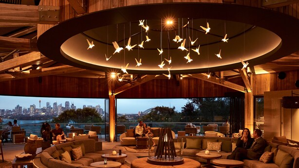 A modern lounge area with a circular seating arrangement, a central fire feature, and a view of a city skyline. - Wildlife Retreat at Taronga (Mosman)