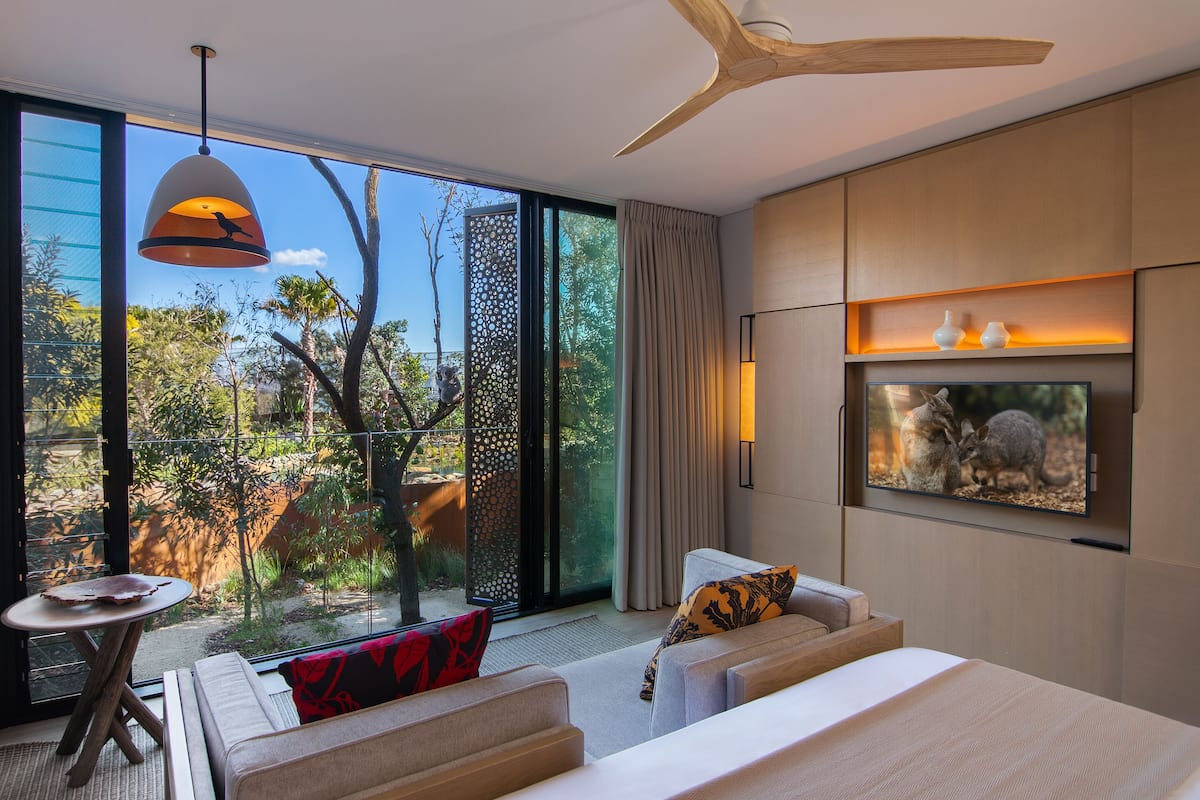 A modern hotel room with a large window, a ceiling fan, a TV, and a sofa with cushions.
