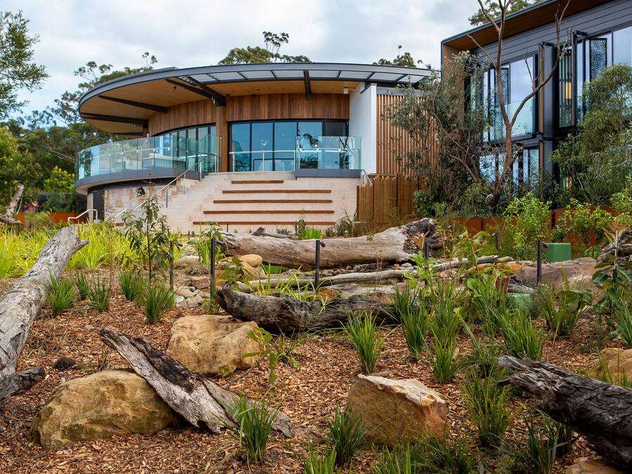 A modern house with a curved glass facade, wooden exterior, and a landscaped garden with rocks and plants.