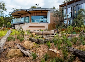 A modern house with a curved glass facade, wooden exterior, and a landscaped garden with rocks and plants. - Wildlife Retreat at Taronga (Mosman)