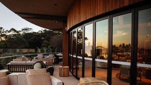 A modern outdoor seating area with wicker furniture and a view of a cityscape through large glass windows. - Wildlife Retreat at Taronga (Mosman)