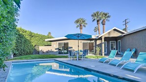 Piscina con sillones de descanso y una mesa, rodeada de una casa y palmeras.