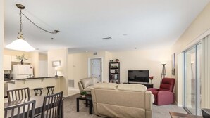 A living room with a sofa, a dining area with a table and chairs, a kitchen with a refrigerator, and a TV in the background. - Minium At Lakeside Golf & Cc - Unit 104 3 Bedroom Apts by RedAwning (Inverness)