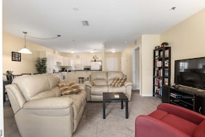 A living room with a beige sofa, a red armchair, a coffee table, and a television on a stand. - Minium At Lakeside Golf & Cc - Unit 104 3 Bedroom Apts by RedAwning (Inverness)