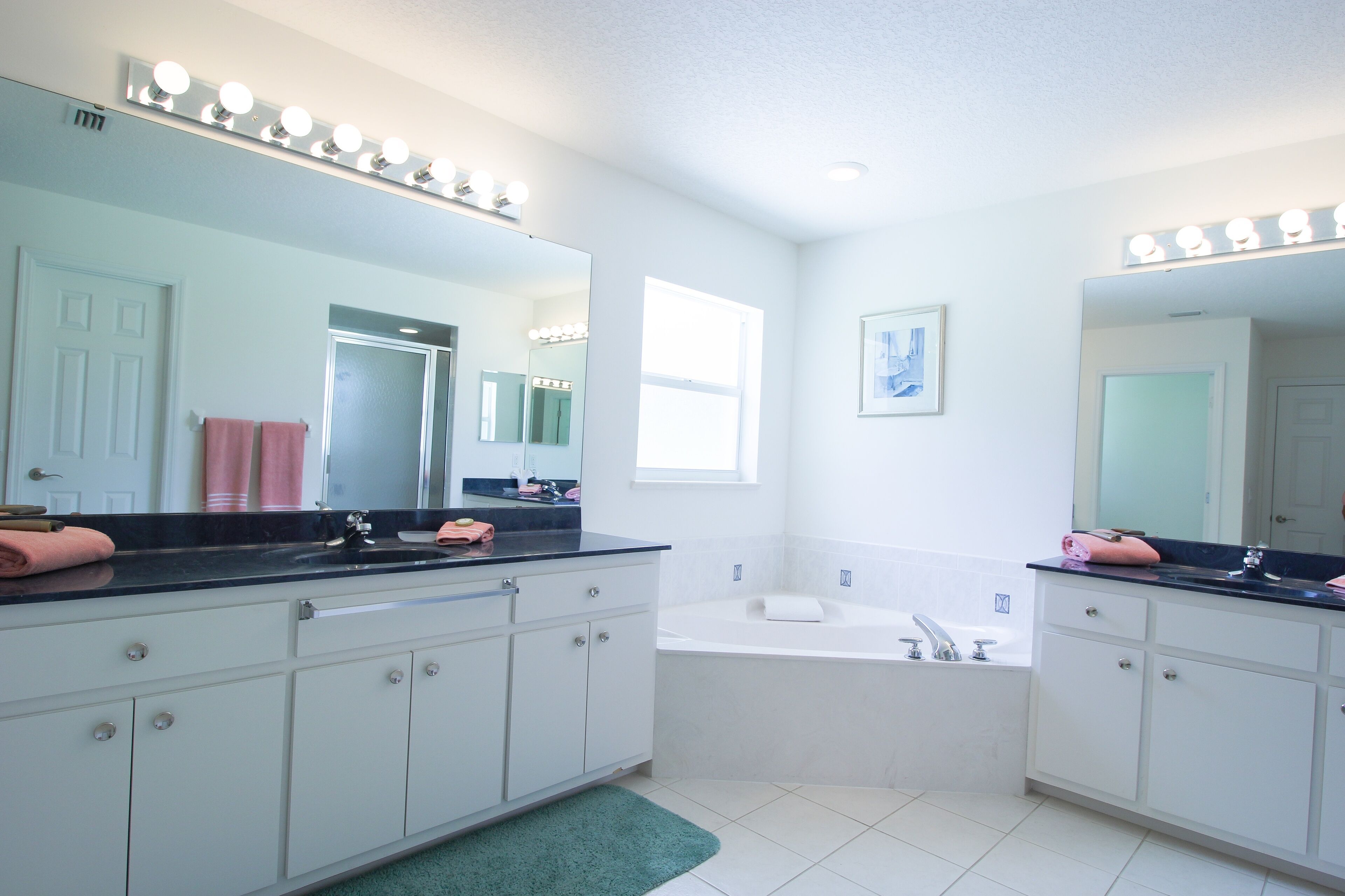 A bathroom with two sinks, a large mirror with lights, a bathtub, and a green rug.