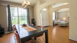 A spacious dining area with a long table and chairs, a large window with curtains, a sofa in the background, and a door leading to another room. - Spacious Modern Pool Home, Family & Golf Trips - 4727 4 Bedroom Home by RedAwning (Inverness)