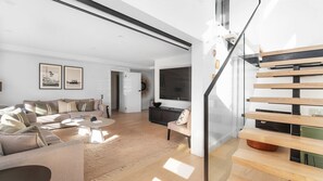 Ein modernes Wohnzimmer mit einer Couch, einem Couchtisch und einem Fernseher. Eine Treppe mit Holzdielen und einer Glastrennwand ist ebenfalls vorhanden.