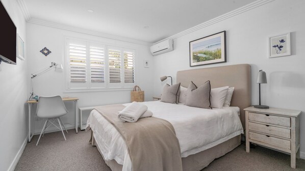 A bedroom with a bed, desk, chair, bedside tables, and a window with blinds.