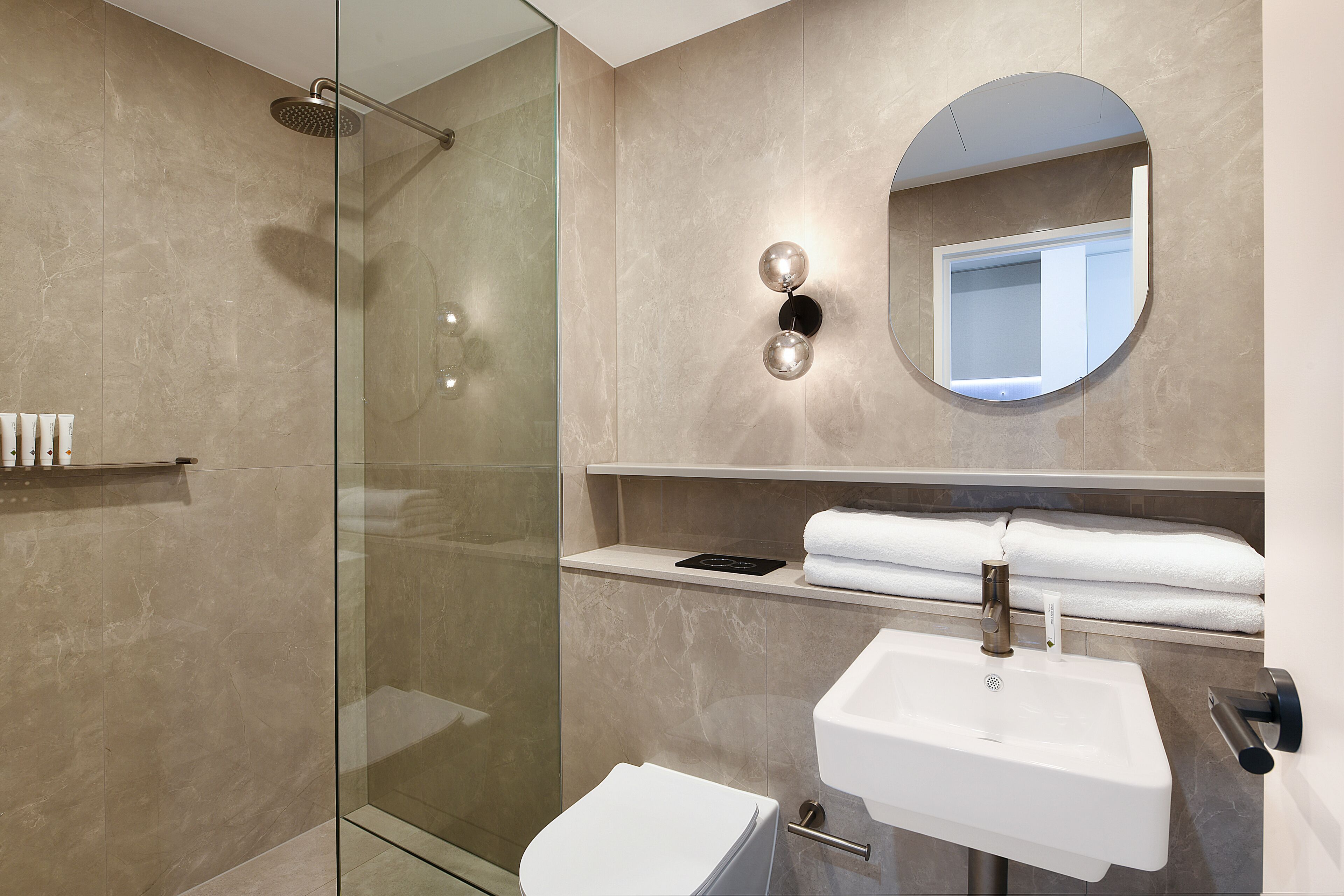 Un baño moderno con ducha envuelta en vidrio, lavabo blanco y espejo redondo.