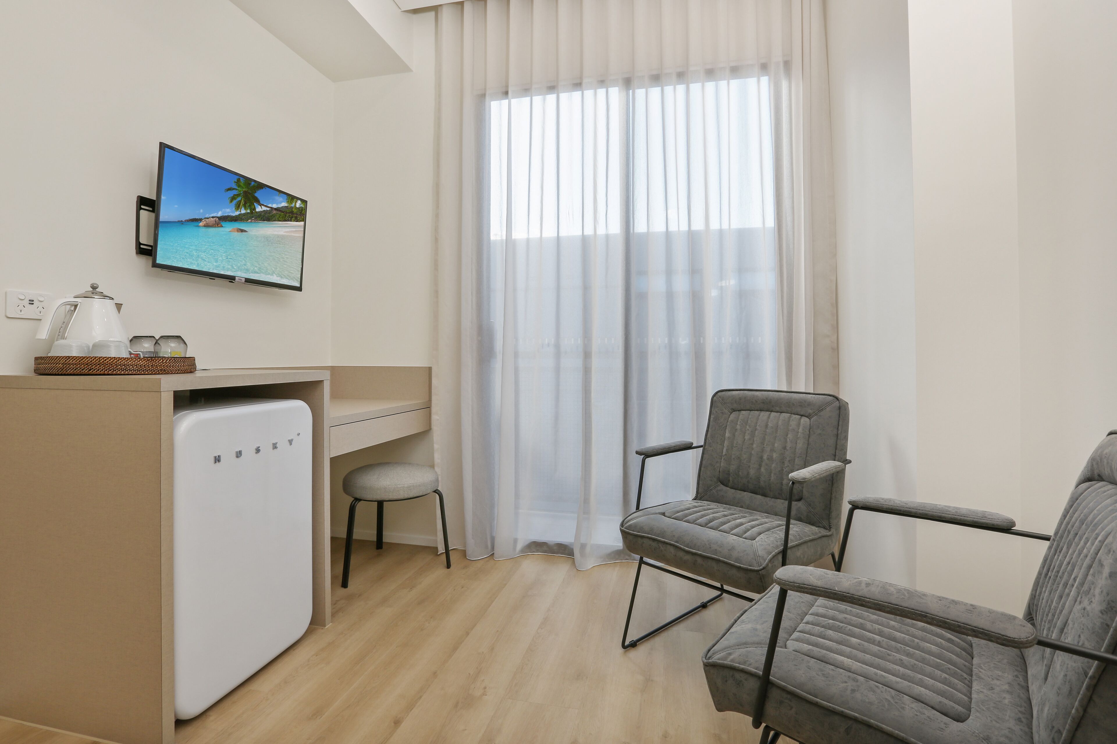 A compact room with a desk, two grey chairs, a small fridge, a TV displaying a beach scene, and sheer curtains.