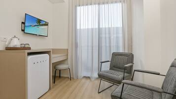 A compact room with a desk, two grey chairs, a small fridge, a TV displaying a beach scene, and sheer curtains.