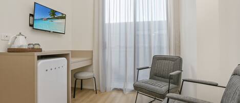 Une pièce de taille modeste, comprenant un bureau, deux chaises grises, un petit réfrigérateur, un téléviseur diffusant une scène de plage et des rideaux légers.