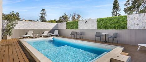 A rectangular swimming pool with a wooden deck, surrounded by lounge chairs and a small table.
