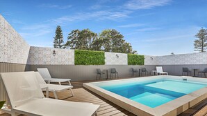 A rectangular swimming pool with a wooden deck, surrounded by white lounge chairs and tables, and a patterned wall with a green hedge. - Nova Kiama (Kiama)