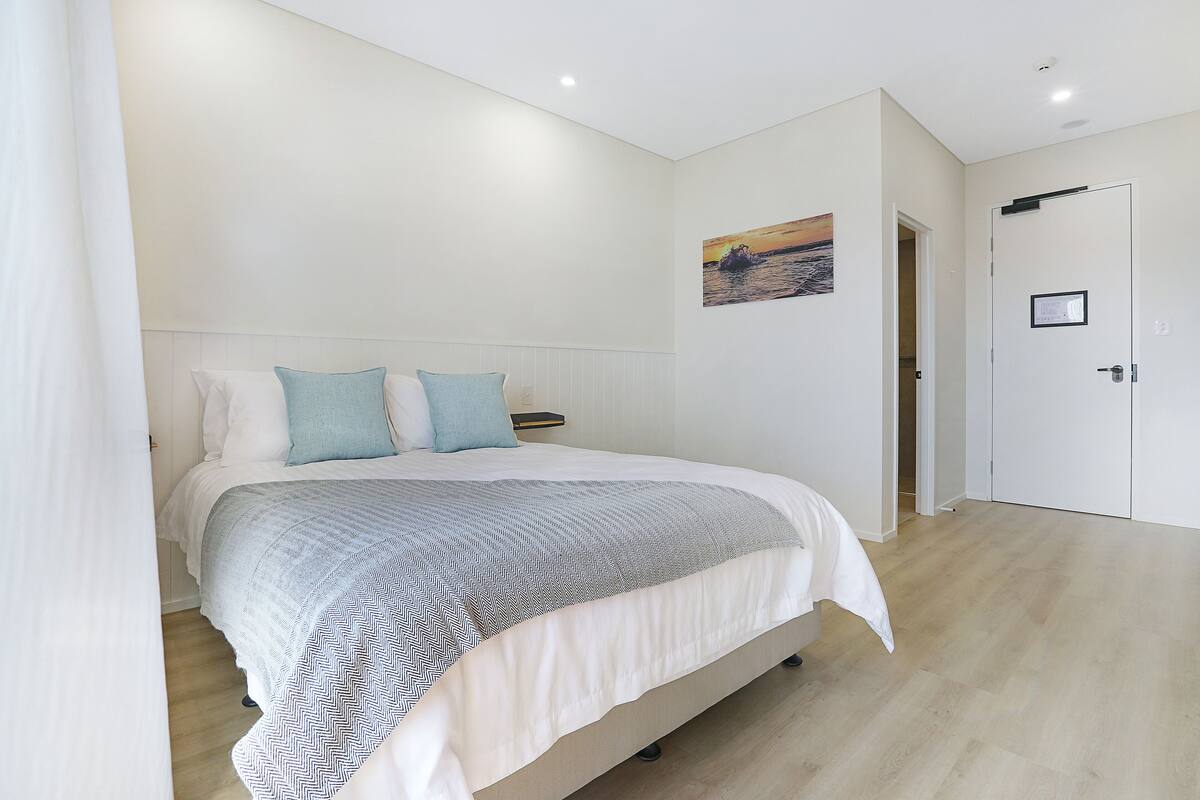 A bedroom with a bed, white walls, a painting, and a door.