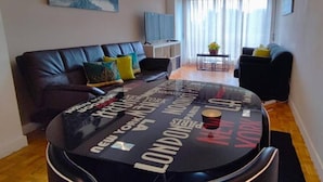 Een moderne woonkamer met een zwarte leren bank, een ronde salontafel met stadsnamen, een televisie en een groot raam met dunne gordijnen.