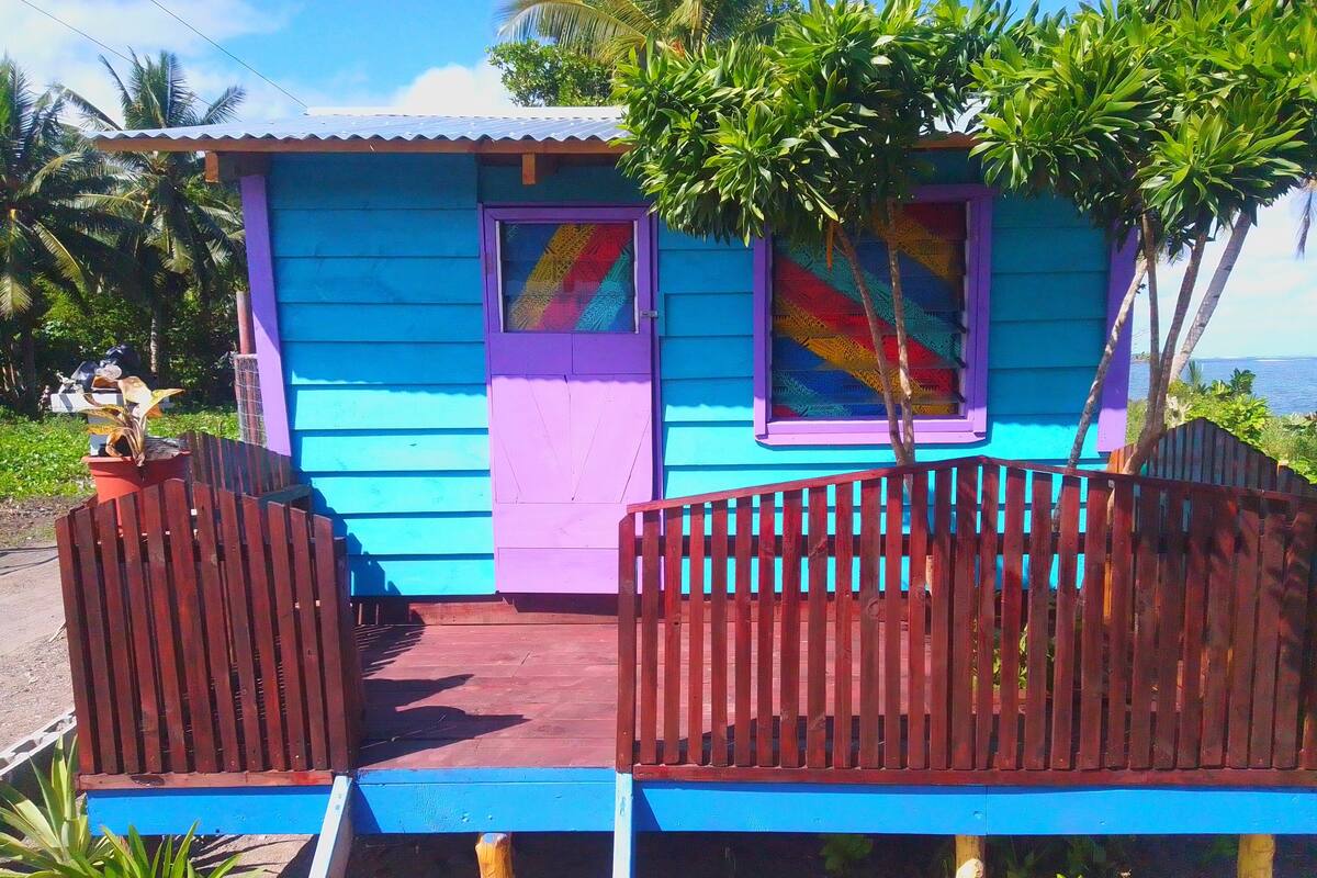 A small, colorful house with a blue exterior and purple accents, featuring a wooden fence and a small porch.
