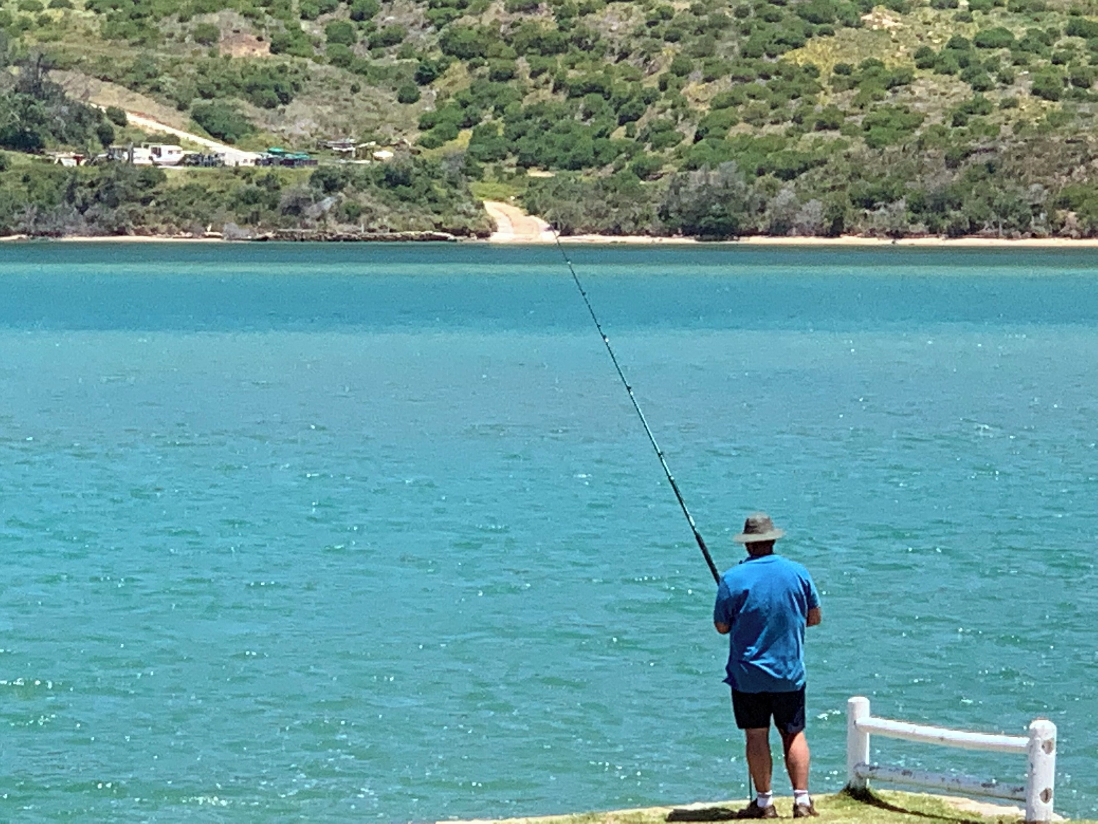 A person fishing by a calm lake with a wooden dock and a white railing.