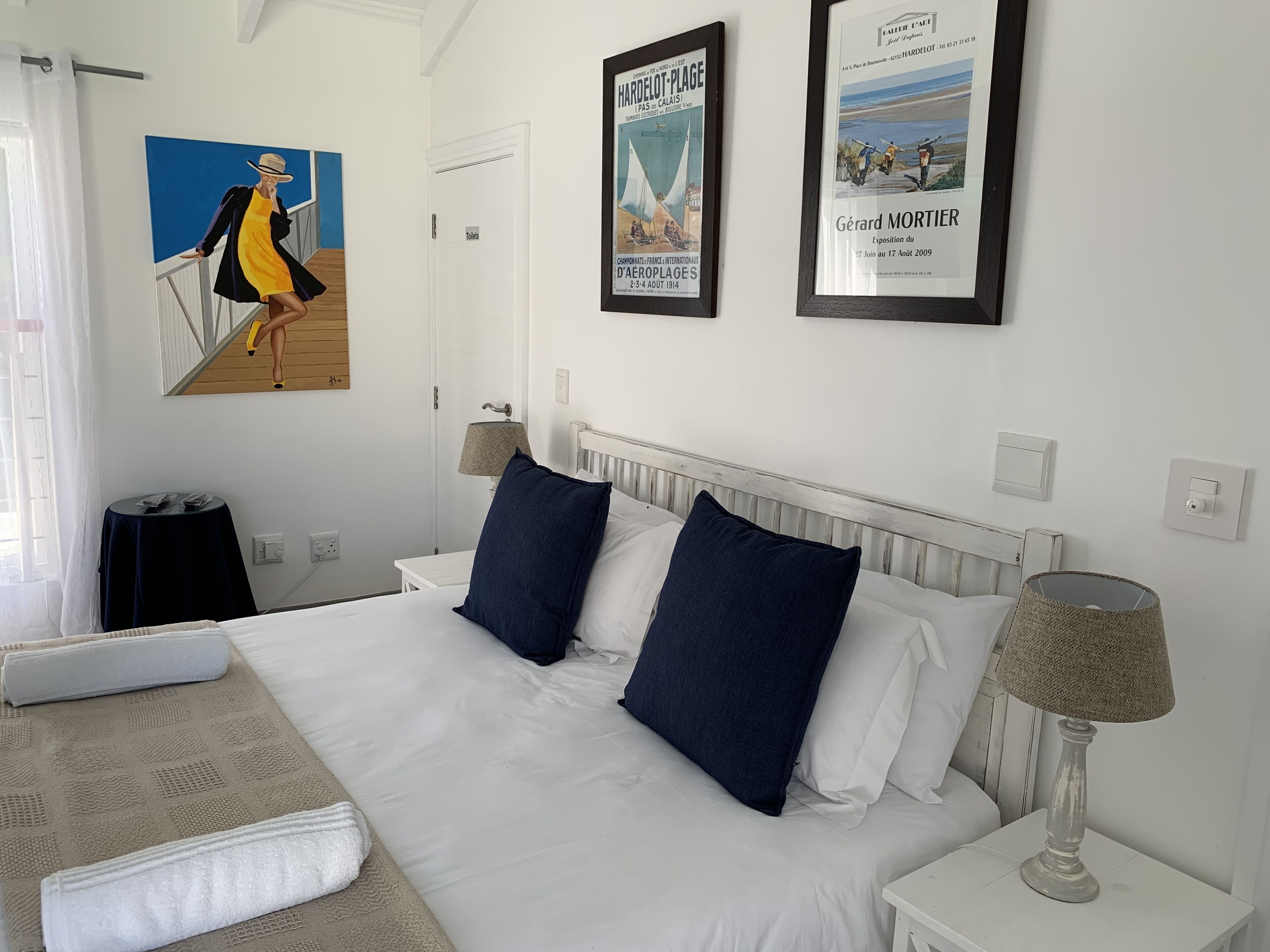 A hotel room with a large bed, two pillows, a nightstand with a lamp, a small table, and a poster on the wall.