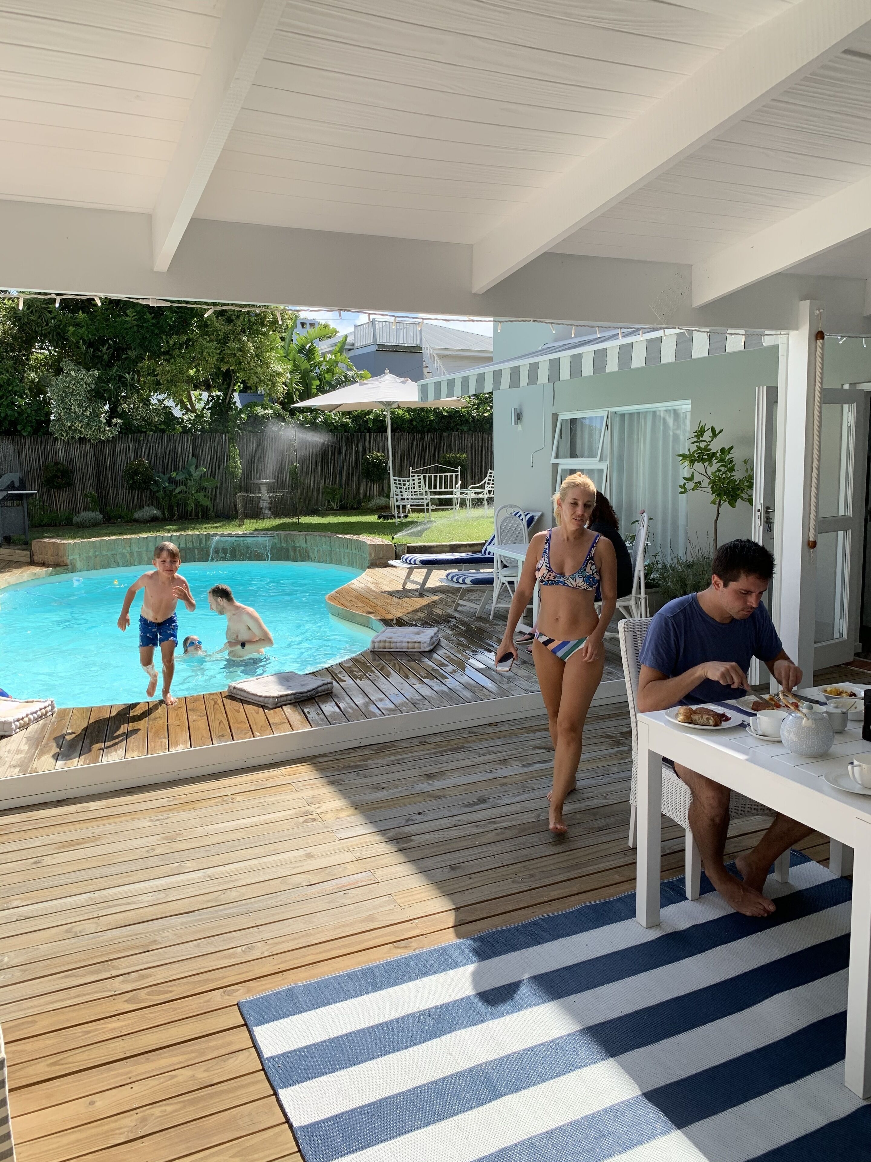 A poolside area with a wooden deck, a dining table with chairs, and a covered seating area.