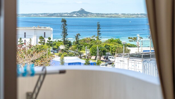 A view from a window with curtains, overlooking a coastal area with buildings, trees, and a clear blue sea. - The Villas Machidoya (Motobu)