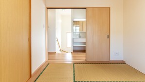 A hallway with a wooden floor, a tatami mat, and a sliding door leading to a room with a mirror and cabinets. - The Villas Machidoya (Motobu)