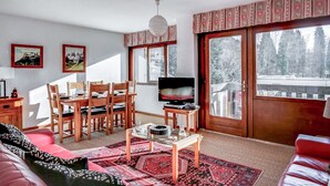 A living room with a dining area, a TV, and large windows with a view of trees.