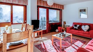 A cozy living room with a red sofa, wooden dining table set, and large windows offering a mountain view.