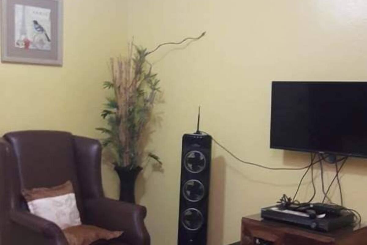 Un salón con un sillón de cuero marrón, una pequeña mesa de centro de madera, un tapete de colores rojo y amarillo, un altavoz negro alto y un televisor sobre un mueble de madera.