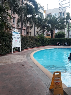 A swimming pool with a caution sign for a wet floor, surrounded by palm trees and a signboard for Avida.