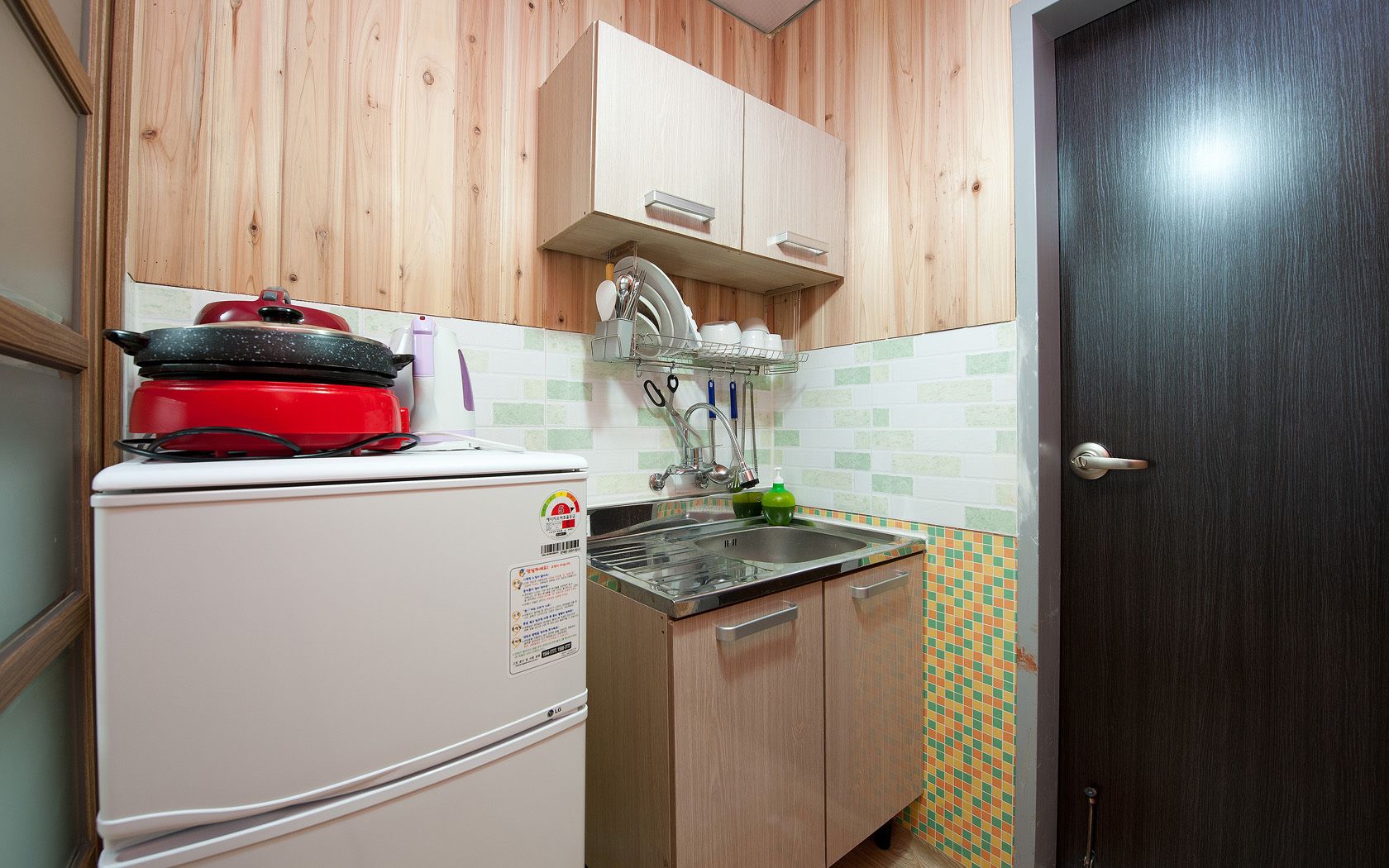 A compact kitchen with a white refrigerator, a red pot on top, a sink with a dish rack, and wooden cabinets.