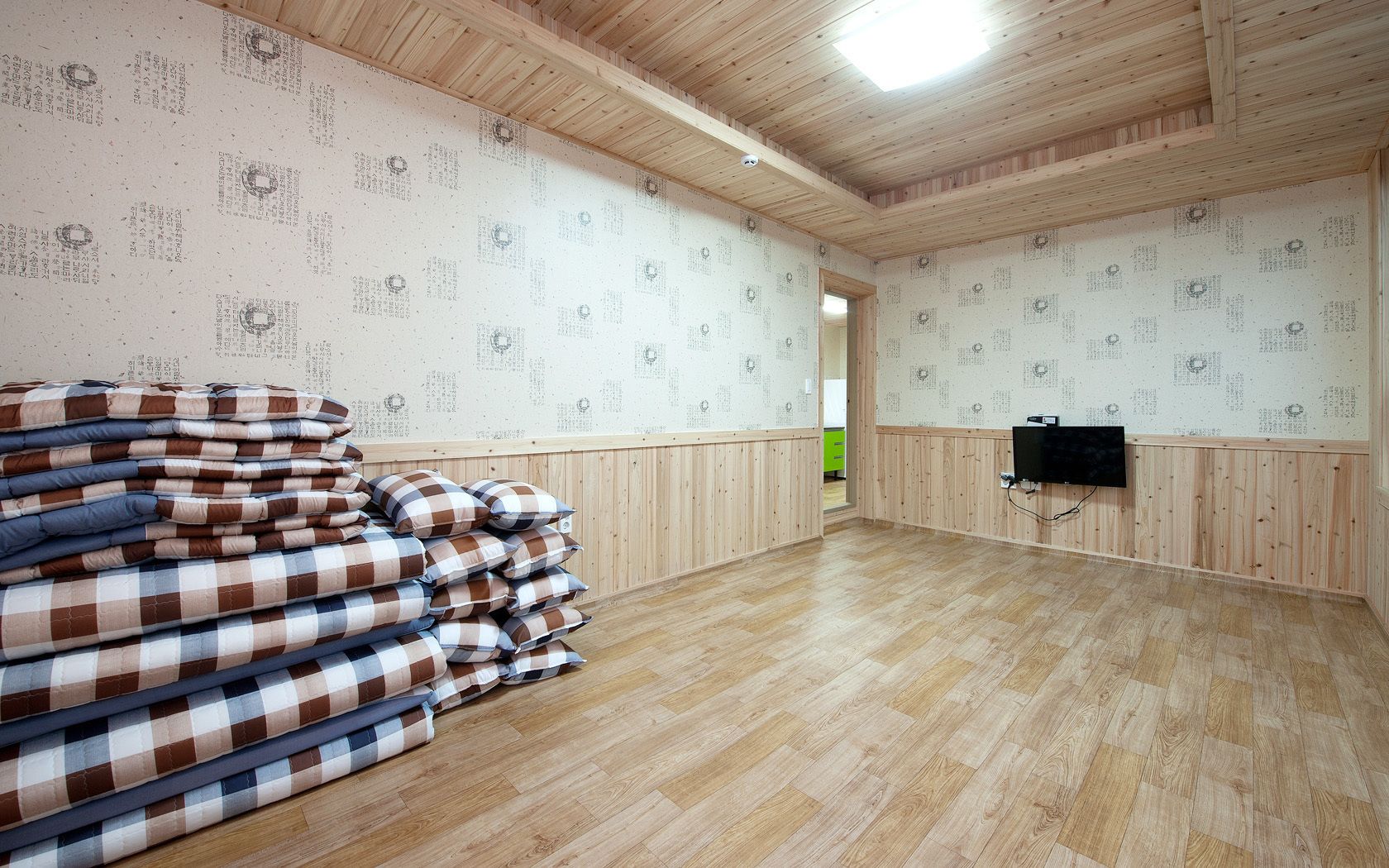 A room with wooden paneling, a television mounted on the wall, and a stack of checkered bedding on the floor.