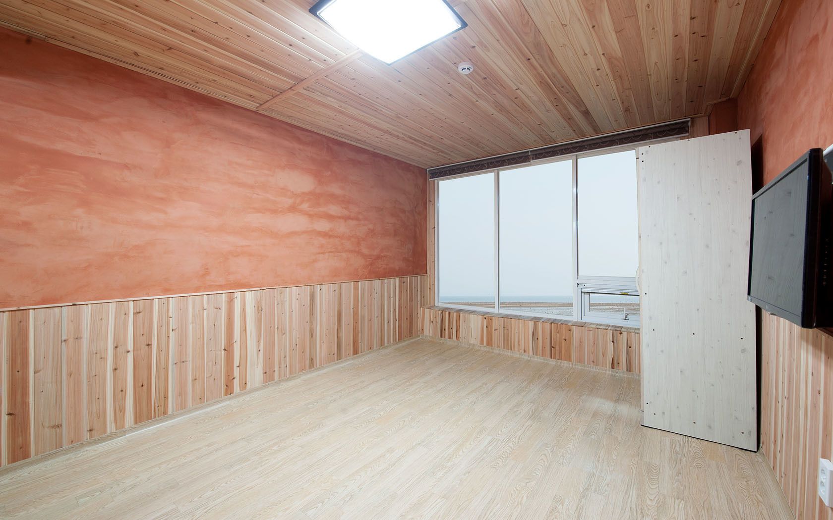 Ein Zimmer mit Holzwänden und -decke, einem großen Fenster und einem an der Wand befestigten Flachbildfernseher.
