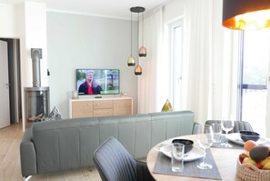 A modern living room with a grey sofa, a dining table set with glasses and plates, a television displaying a program, and a fireplace.