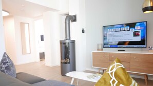 A modern living room with a grey sofa, a white coffee table, a TV displaying a news channel, a wood-burning stove, and a wooden cabinet.