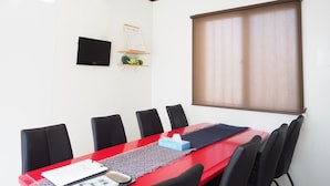 A conference room with a red table, black chairs, a mounted TV, a whiteboard, and a window with blinds. - Holiday Homes TSUBOYA 2nd (Naha)